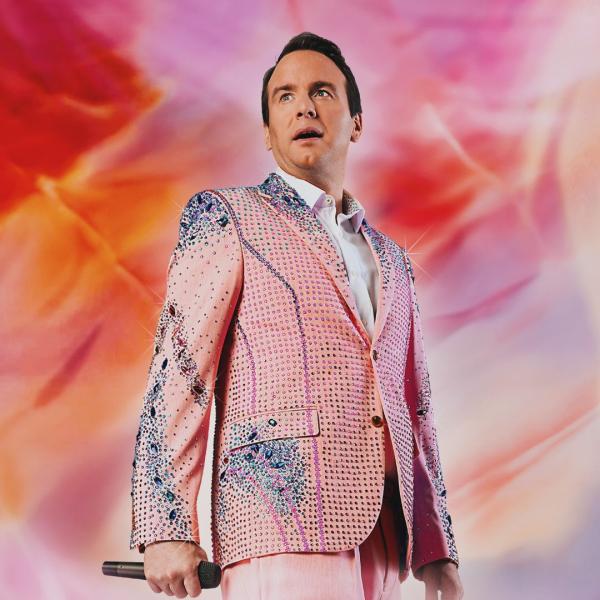 Matt Forde wearing a pale pink bejewelled suit with blue details and white shirt underneath. He is holding a microphone down by his right side and is against a pink and orange wispy background. The image is mimicking Taylor Swift's Eras Tour artwork.