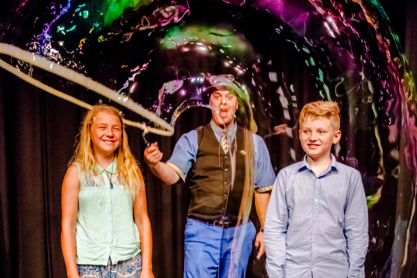 Two children stand either side of a man with a large bubble wand, he is producing a large tube shaped bubble to cover them both.