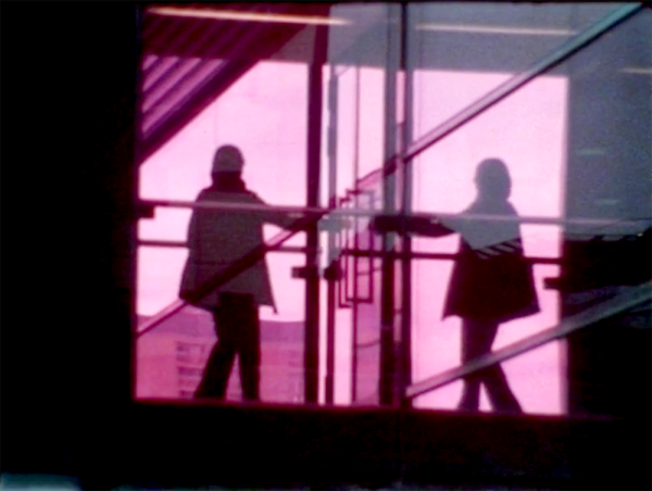 Two people (or a reflection) behind a window in pink light standing on either side of a door.