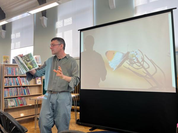 Simon, a white man with dark hair and glasses, holds up a book in his right hand and gestures with his left hand open. He is mid-speech. Behind him to the left are rows of library bookshelves, and to the right is a projector screen showing an image of a butterfly.