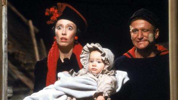A woman in a red hat and sarcf holds a baby in old fashioned clothes with a man in sailor clothing next to her smoking a clay pipe