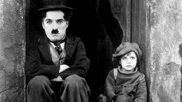 Charlie Chaplin, smartly dressed and arms crossed, sits on the step of a building next to a small boy.