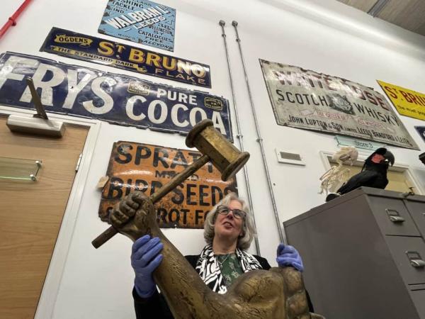 A picture of Curator of Leeds and Social History Kitty Ross holding a goldbeaters sign (arm and hammer) in front of adverts for Fry's Cocoa, Bird Seed etc.