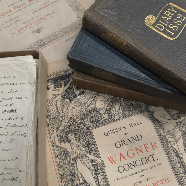 Various papers, notes and books about music with a pile of bound volumes and an ornately illustrated poster which reads "Queens Hall Grand Wagner Concert", all laid out on a table