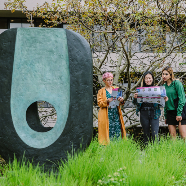 Three people holding public art maps beside a blue and black oval sculpture with a hole through it