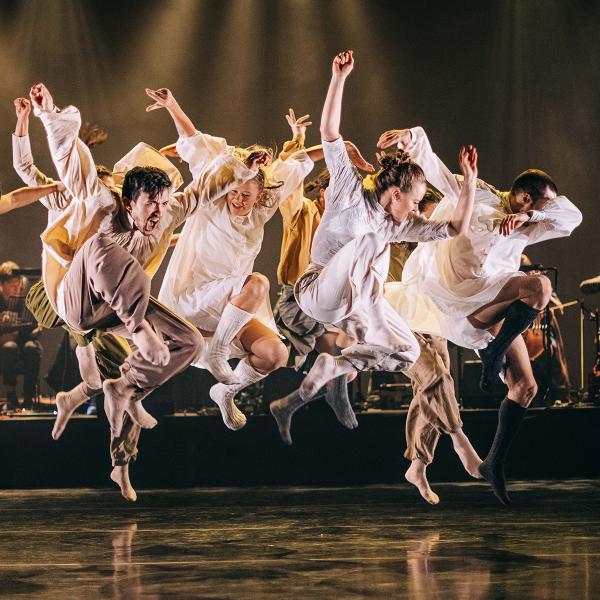 Group of dancers jumping in the air, with folk band behind on stage