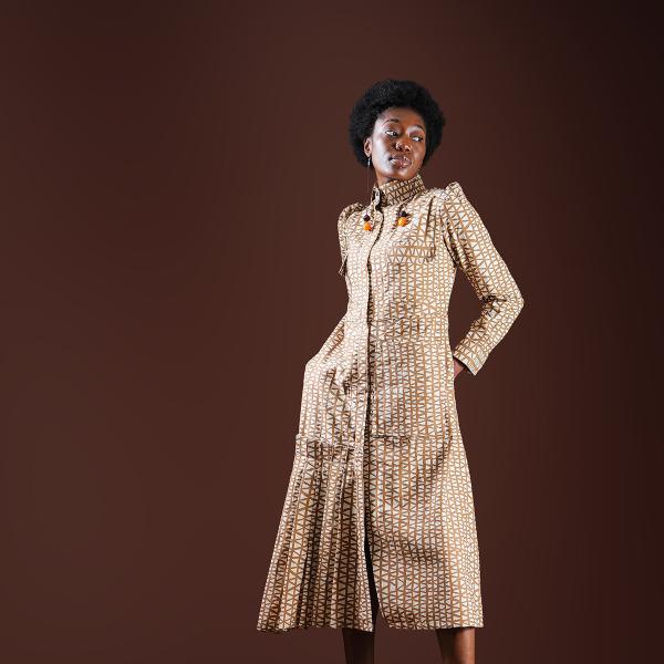 A black woman poses wearing cream patterned dress in from of brown background