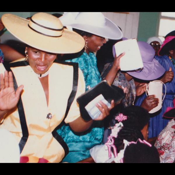 Women in a selection of church hats raise their hand holding their purses