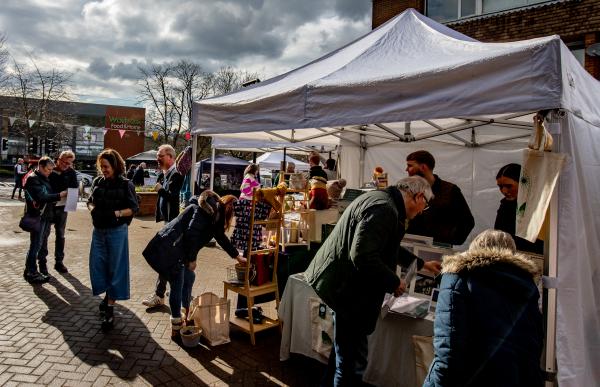The photo shows an autumnal market day with people looking at a market stall and standing together chatting.