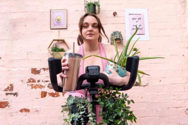Lydia in milennial pink on a spin bike clutches a smoothie and a pot plant