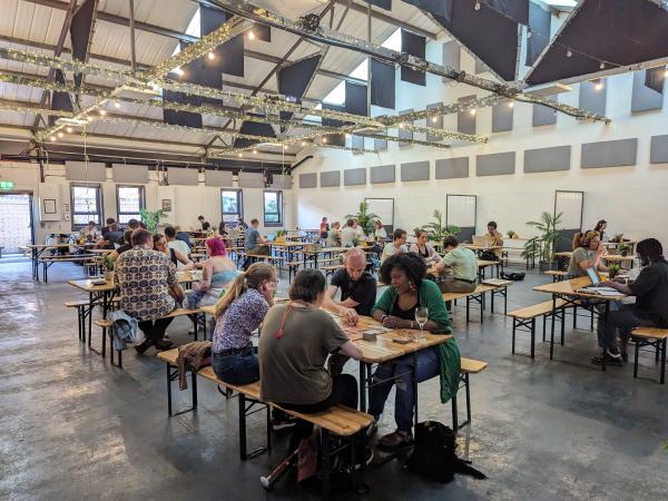 A large room with a high ceiling and fairy lights high up. People are sitting on benches at tables and playing boardgames.
