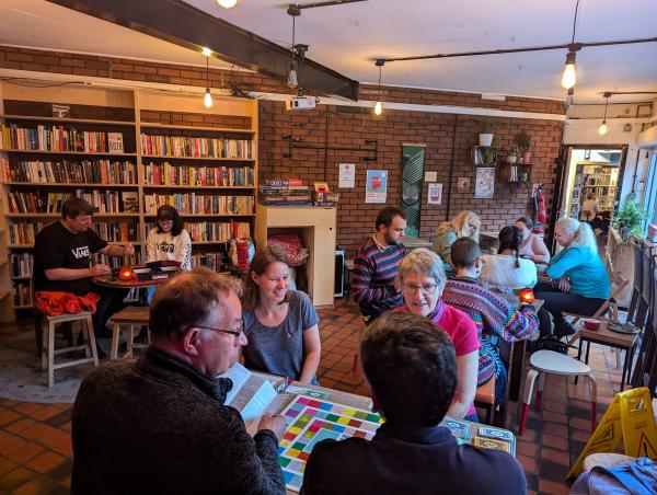 A room with full book shelves along one wall. People are sitting at tables in groups and playing games.