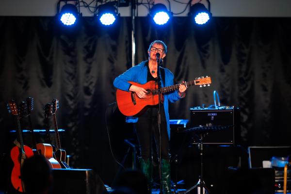 Older woman stood on a stage with a blue jacket on. She's playing the guitar and singing into a microphone.