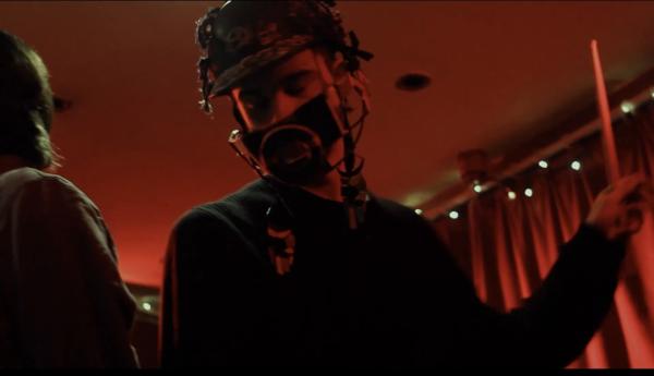Herbie Hopkins (male, black jumper, holding drumstick with left hand) wearing a helmet decorated with charms and drawings which also covers his mouth in a red-lit room with fairy lights and red curtains