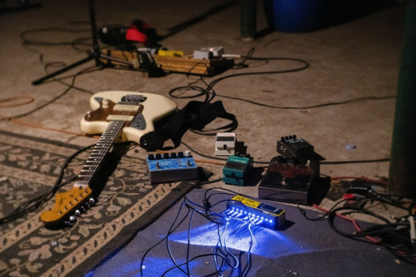 shot of various guitars, pedals and audio cables on a rug and floor. Blue lights shine from one of them. Signs of a jam session