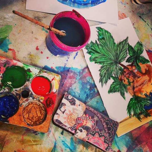 A photograph of a paint pallet with blue, green, red and gold paint, a water pot and a painting of a plant. 