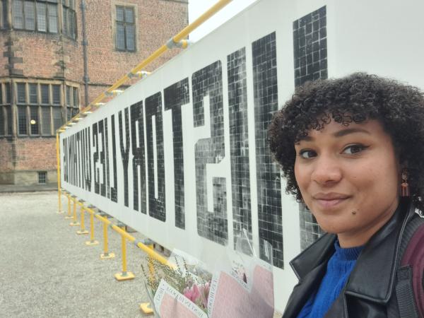 A women with dark curly hair stands in front of a big poster