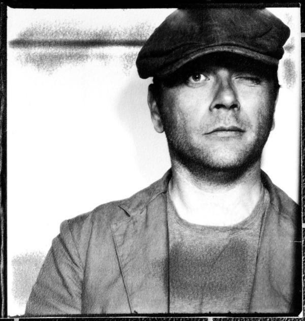 Black and white close u photograph of John Angerson. John is wearing a dark 'baker boy' style cap and is winking at the camera with a slightly startled expression
