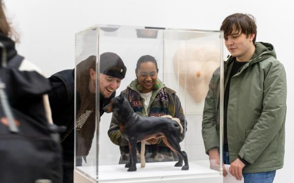 A group of three people stand around a sculpture of a dog, peering at details and laughing as they discuss the work.