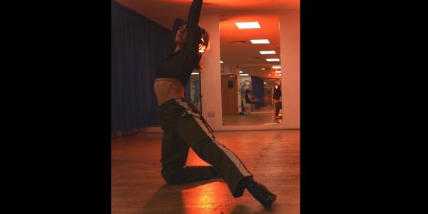 Solo female dancer in a studio, with orange lighting. She is wearing green tracksuit trousers with a white strip down the outside centre of the leg,a green long sleeved cropped top and black lace heels. She is positioned on her knee with her left leg out straight ad arms up, one placed on here head.