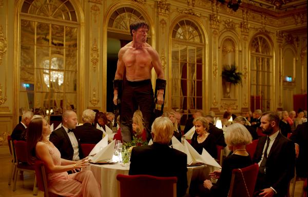 image shows man with no top on stood on a tabel in a formal room surrounded by people wearing formal attire