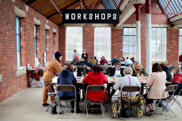 kim stands at the erhad of a table in an old indusrial space. people are sat around the table a 'workshop' sign hangs above their head.