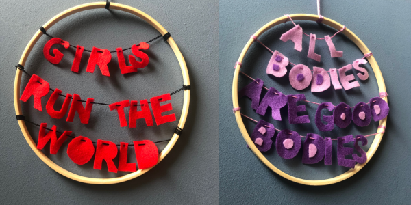 Image of two IWD wall art pieces made from felt inside a wreath. The first one says 'Girls Run The World' in red and the second says 'All Bodies Are Good Bodies' in pink and purple felt.