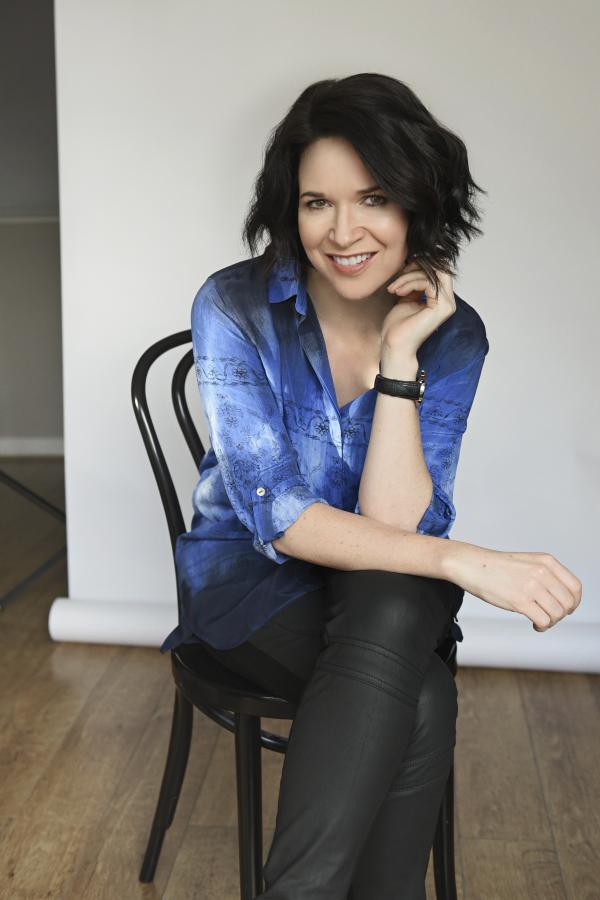 Image of author sitting on chair with left leg crossed over right. Looking into camera, wearing dark trousers and blue shirt