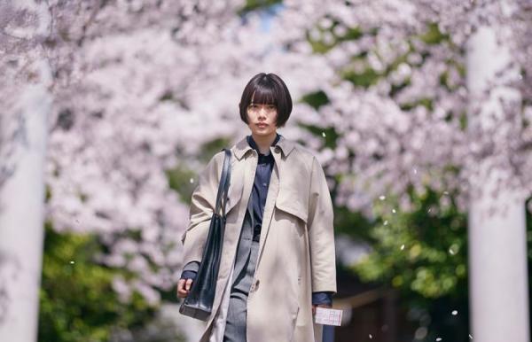 Image shows daytime, a Japanese woman is walking outside. She is cropped below the knee and in the background we see two cherry blossom trees.