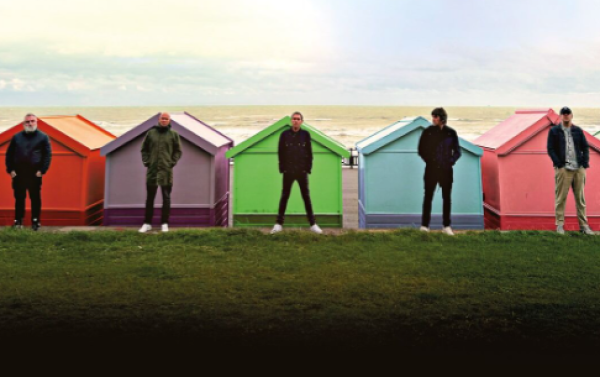Image of the band lined up in front of very colourful seaside sheds with the beach and sea in the background and a patch of green grass in front of them. 