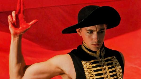 Man dressed in military waist coats and black wide rim hat is holding up hand that's covered in red, Infront of a red background.