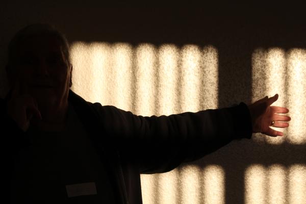 a photograph showing the silhouette of an older man standing with one arm outstreched with the shadow of a venitian blind behind him.