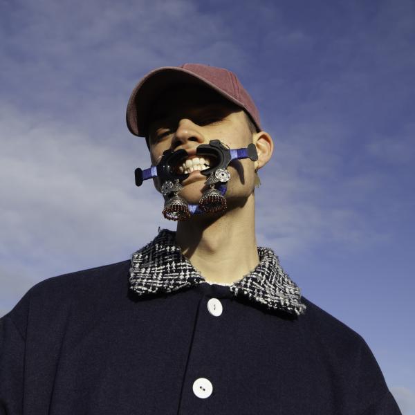 Portraits of Herbie Hopkins in front of a near clear sky with a cloud. Wearing baseball cap and shirt with plaid collar. His pose involves him bearing his teeth, but by what appears to be a dental device to push his lips away from all his teeth
