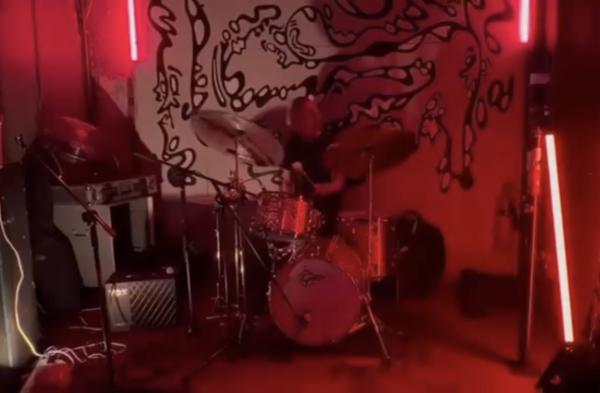 A drum kit in the middle of the room played by Oli Reeves infront of a wall with paintings of distorted faces.