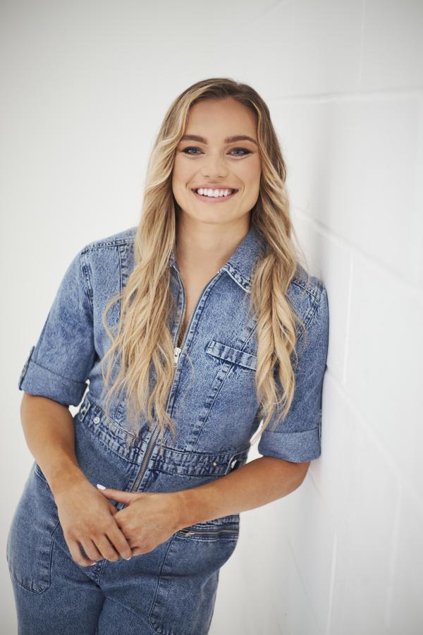 Picture of Jodie wearing a denim blue jumpsuit leaning against a blank white wall smiling