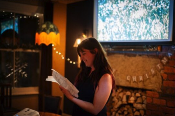 warm interior showing a woman reading aloud at a pub or venue, from a sheet. Image is focused just on her.