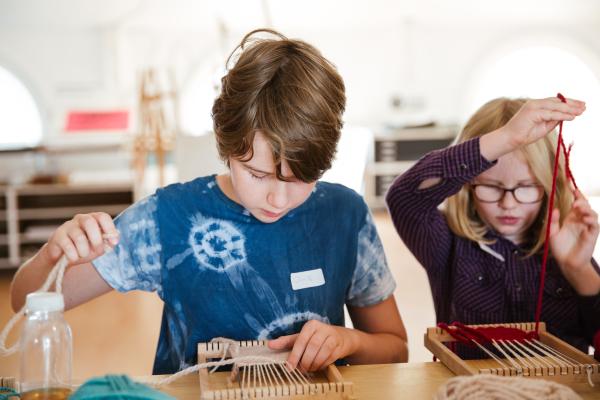 TWO YOUNG PEOPLE WEAVING ON TAPESTRY LOOMS AT A TABLE WITH COLOURFUL YARN