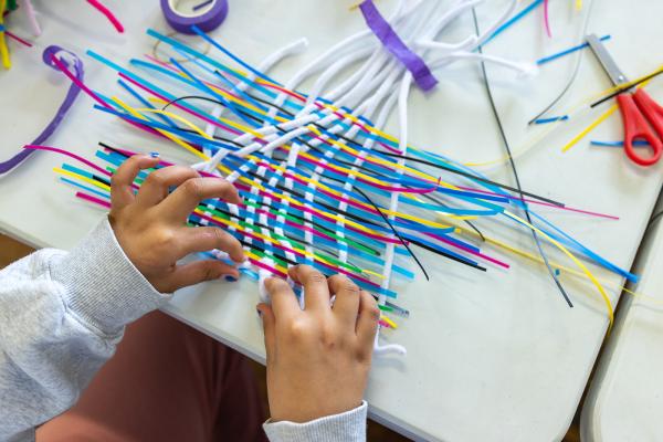 Young persons hands weaving brightly coloured paper strips and pipe cleaners on a table