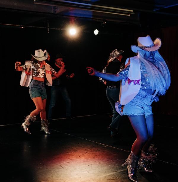 Two women dancing and having a good time at Decodance, dressed in cowgirl waistcoats, hats and boots. Mid stomp, gleeful expressions