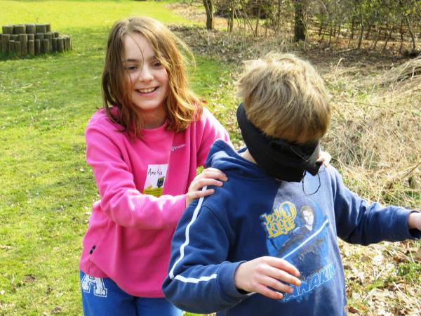 A girl wearing a pink top guiding a blindfolded boy wearing a blue top