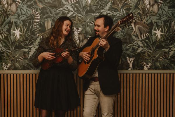 Karoliine-Lisette Kõiv (fiddle and vocals) and Stuart Ironside (guitar and vocals) stand with their instruments, smiling and looking at each other, in front of a decorated wall.