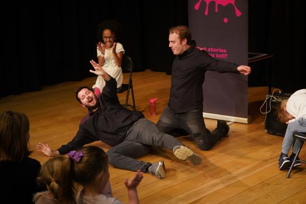 two men roll flamboyantly on the stage floor while children clap and laugh delightedly