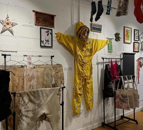 Rails of natural dyed clothes on the left and right, yellow, embroidered beekeeping suit hung in the middle surrounded by framed contributions by members of the group