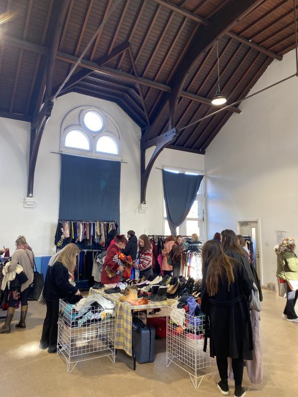 A group of people rummaging through baskets and rails of clothes in a community centre hall with high wooden ceilings.
