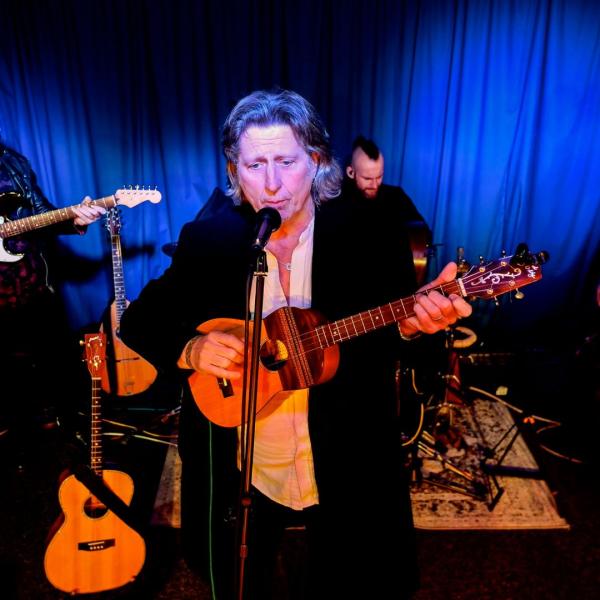 Steve Knightley playing a tenor ukulele and singing into a microphone on a stand with a band behind him.