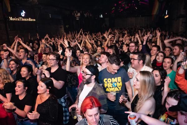 A large, excited crowd of musical theatre fans cheering, dancing, and singing along in a packed nightclub. Many are smiling, clapping, and wearing themed outfits, including a 'Come From Away' t-shirt. The atmosphere is lively, joyful, and full of energy, with colourful lighting and a sign reading 'Musical Theatre Raves' in the background