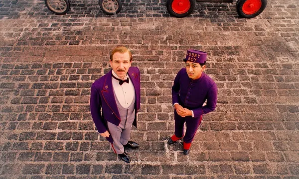 Ralph Fiennes and Tony Revolori, dressed in the Grand Budapest Hotel's purple livery, look upwards with questioning expressions.