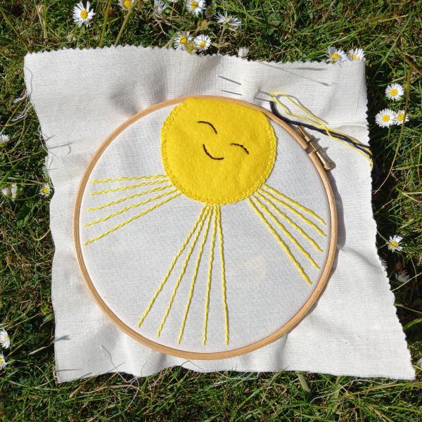 An embroidery hoop with some plain coloured fabric which has been embroidered with a yellow sunshine which has a smile and closed eyes.