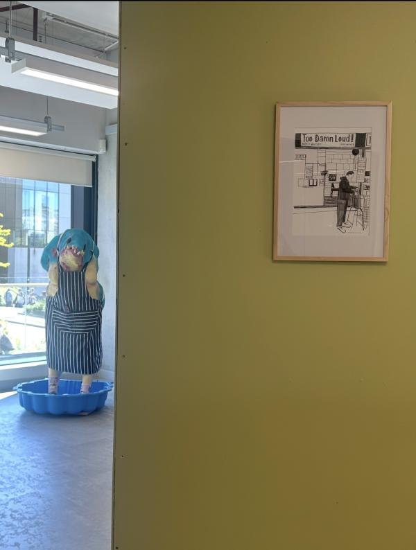 A gallery space with a green wall that has a framed drawing of a music shop and a sculpture of a creature in the space to the left.