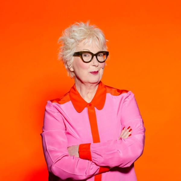 Jenny Eclair with her arms crossed wearing a pink and orange shirt and her signature black glasses against an orange background.